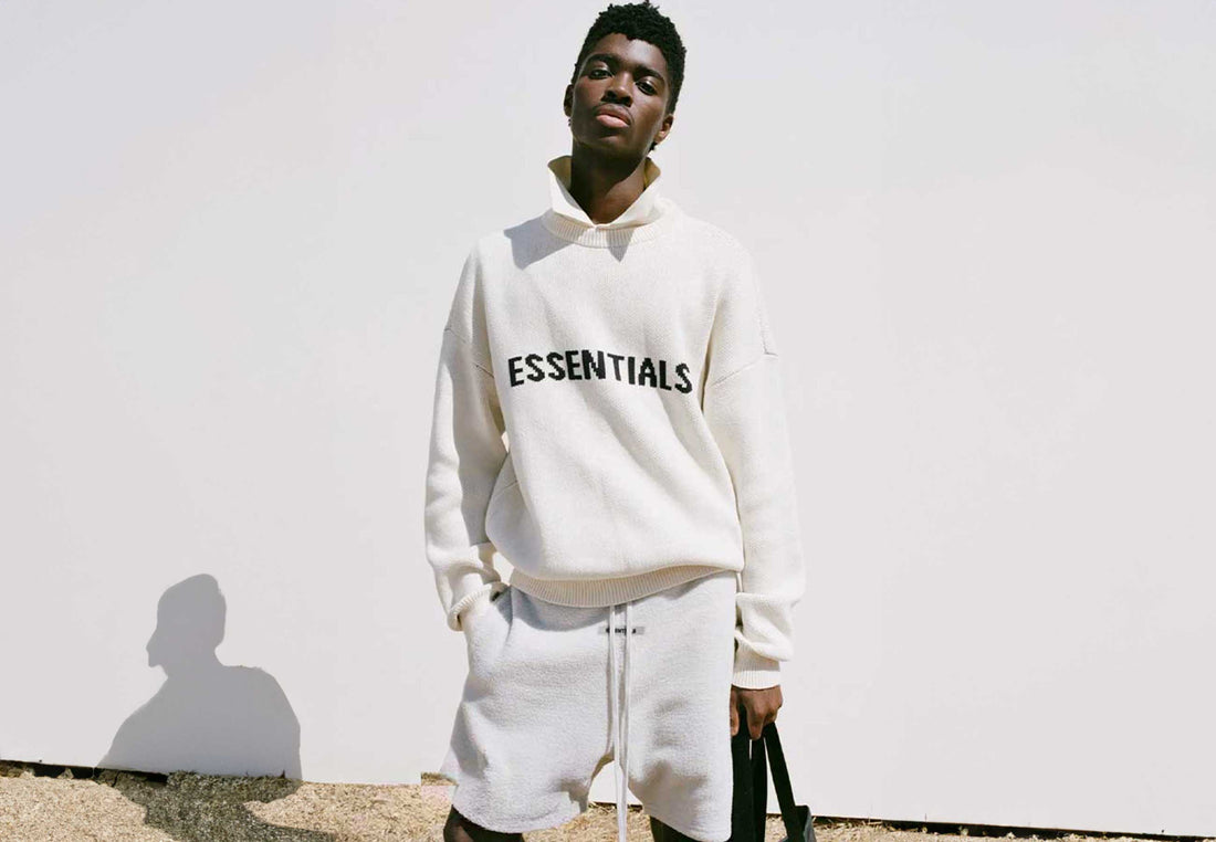 Image depicted male model facing camera wearing an Essentials hoodie, Essential shorts, and Essentials tote bag.