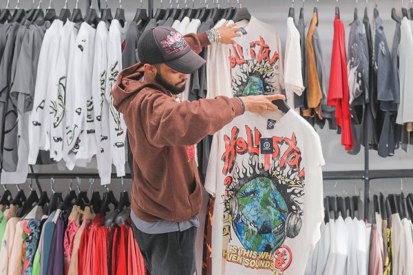Image show inside the Restock AR Store Front. In this image, the employee is organizing the clothing racks and is holding up a Hellstar Studios Heaven On Earth Cream T-Shirt.
