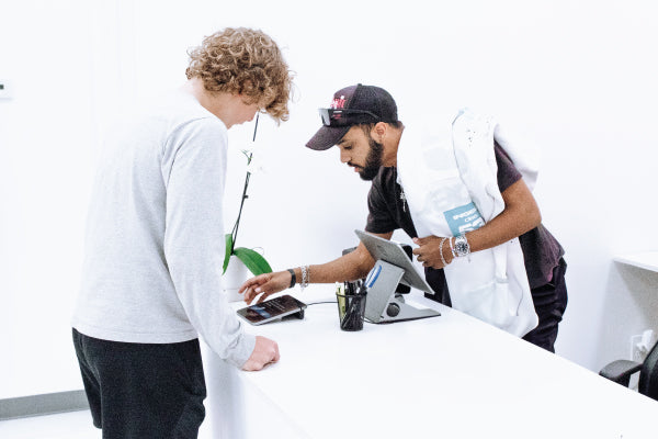 Image shows customer being helped by employee in-store. Customer is checking out, and buying a Sp5der Worldwide Websuite White Hoodie.