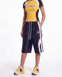 A model wearing a yellow, black, and white athletic top and matching black athletic shorts with white stripes and drawstrings, paired with yellow and white sneakers, against a plain white background.