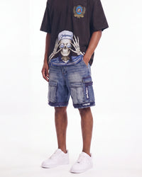 A model wearing blue denim cargo shorts with a large graphic print of a skull wearing a cap on the front, paired with a black t-shirt and white sneakers, against a plain white background.