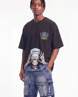 A man wearing a dark brown oversized graphic t-shirt with a large skeleton print and a chest patch, paired with blue denim cargo shorts against a plain white background.