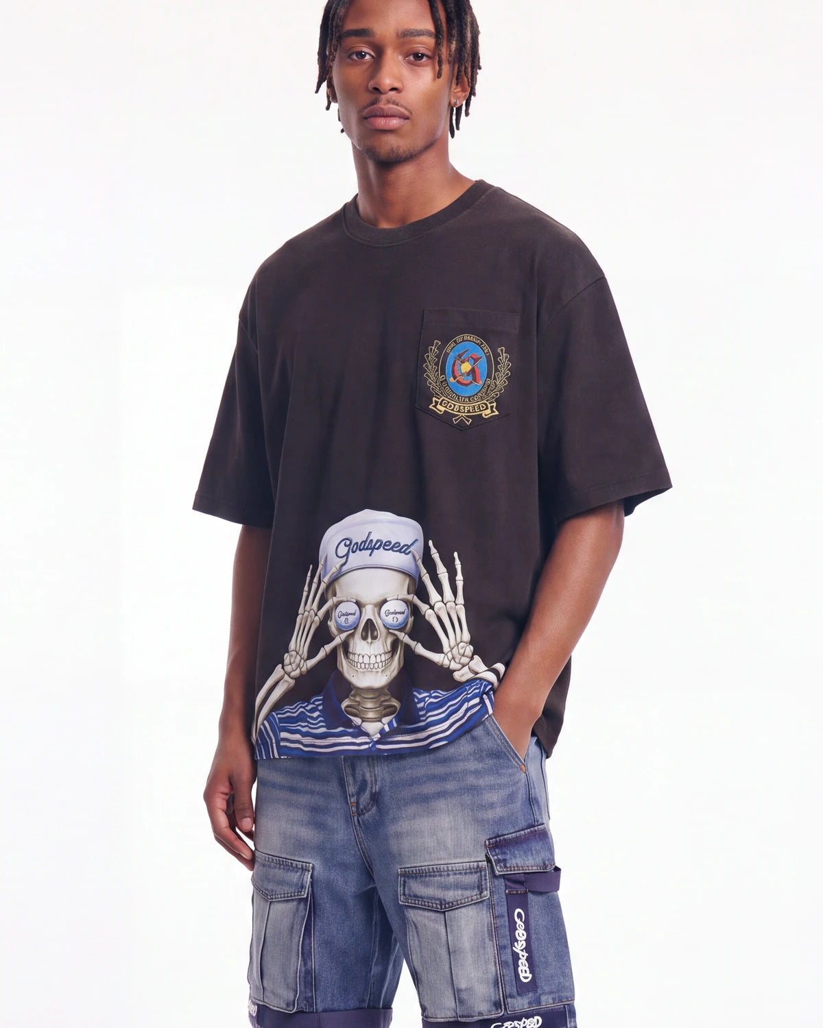 A man wearing a dark brown oversized graphic t-shirt with a large skeleton print and a chest patch, paired with blue denim cargo shorts against a plain white background.