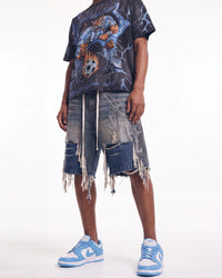 A model wearing a dark graphic t-shirt, heavily distressed denim shorts with frayed edges, and light blue and white sneakers, photographed against a plain white studio background.