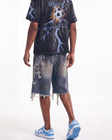 A person wearing a black graphic t-shirt with a soccer ball and lightning print, distressed denim shorts with frayed hems, and blue and white sneakers, shown from the back against a white studio background.