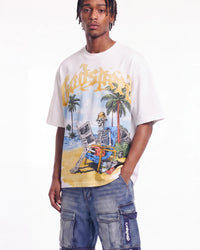 A man wearing a white graphic t-shirt with a colorful beach scene featuring a skeleton in a chair and the word 'Vendetta' in yellow, paired with blue denim cargo shorts.