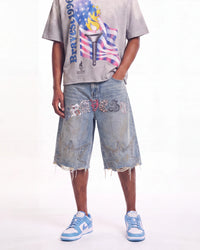 A person modeling light-wash distressed denim shorts with embroidered patches and frayed hems, paired with a graphic t-shirt and light blue sneakers, against a plain white studio background.