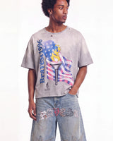 A man wearing a faded light gray graphic t-shirt with a distressed design featuring the word 'Braves' and an American flag motif, paired with light-wash jeans.