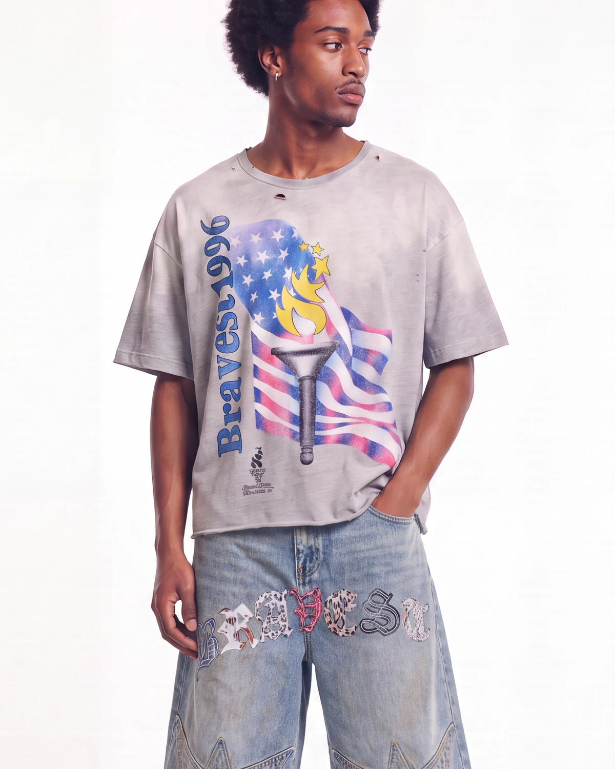 A man wearing a faded light gray graphic t-shirt with a distressed design featuring the word 'Braves' and an American flag motif, paired with light-wash jeans.