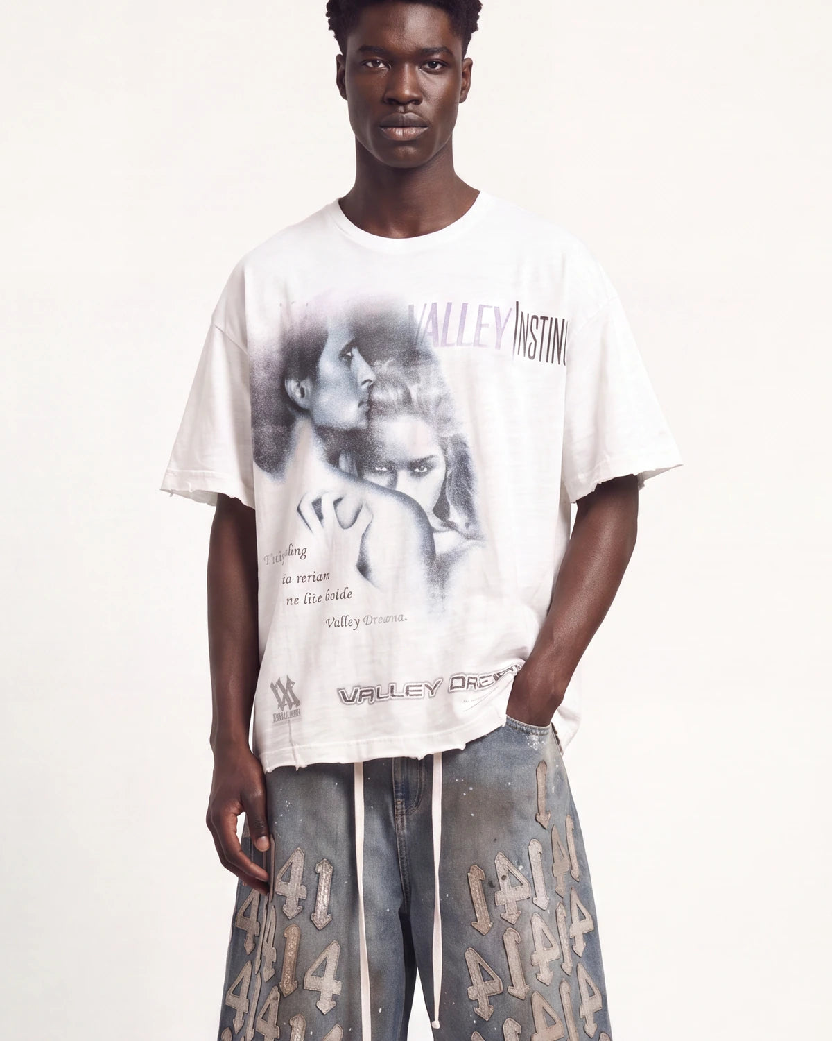 A male model wearing an oversized white graphic t-shirt with a black and gray print of a couple and the text 'VALLEY INSTITUTION', paired with gray patterned shorts, against a plain off-white background.