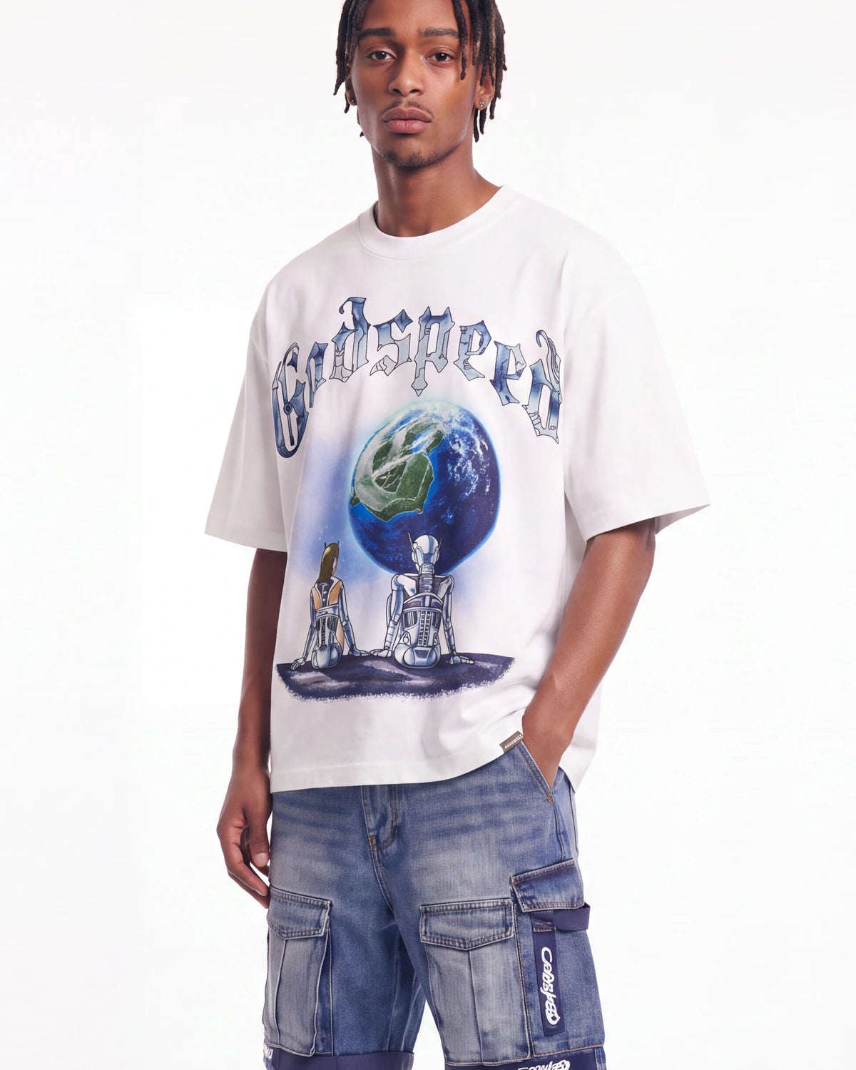 A man wearing a white oversized t-shirt with a graphic print of the word 'Godspeed' above a globe and two seated figures, paired with blue denim cargo shorts, against a plain white background.