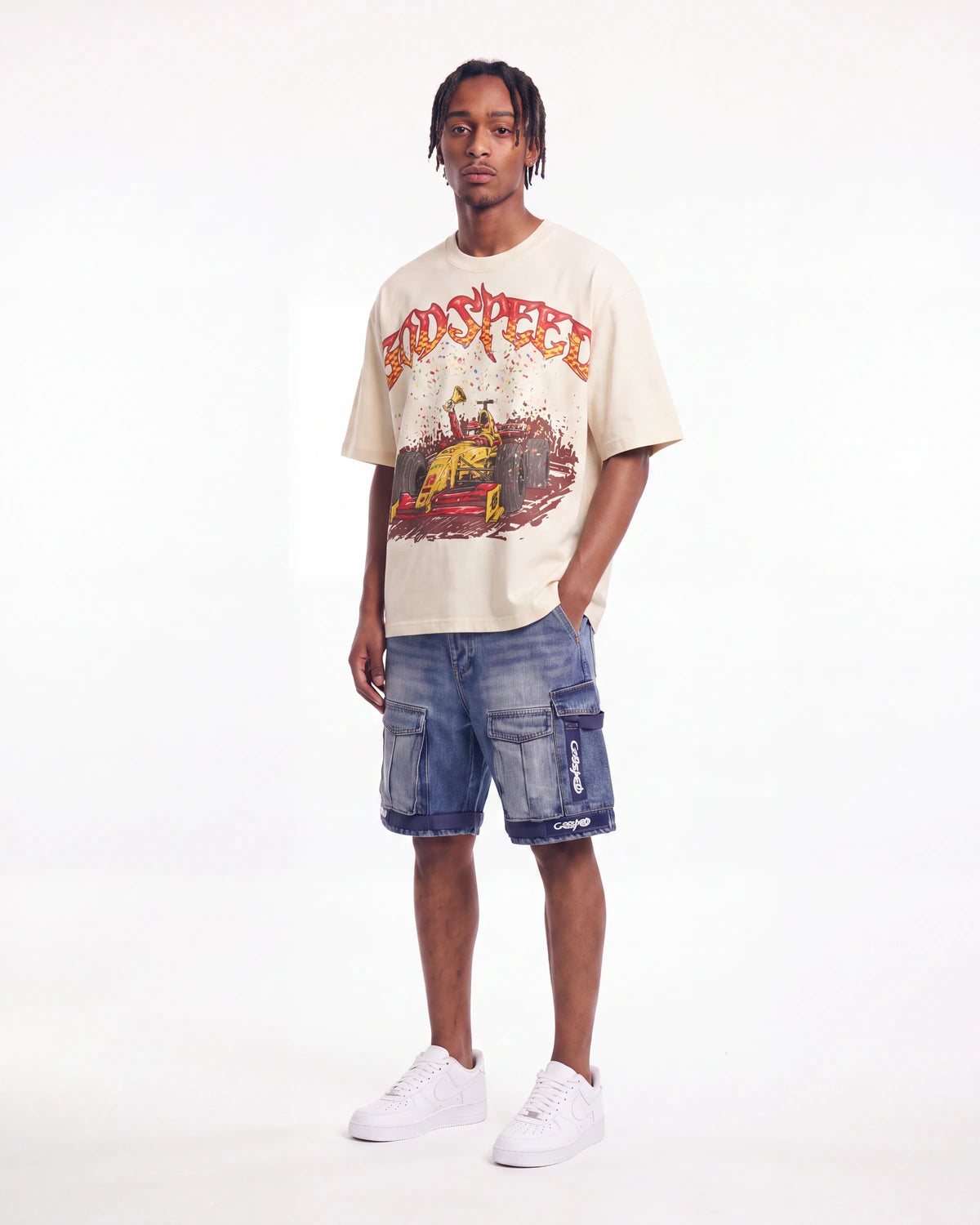 A man modeling a beige graphic t-shirt with a red and yellow 'TOO SLEEP' design, paired with blue denim cargo shorts and white sneakers, against a plain white studio background.