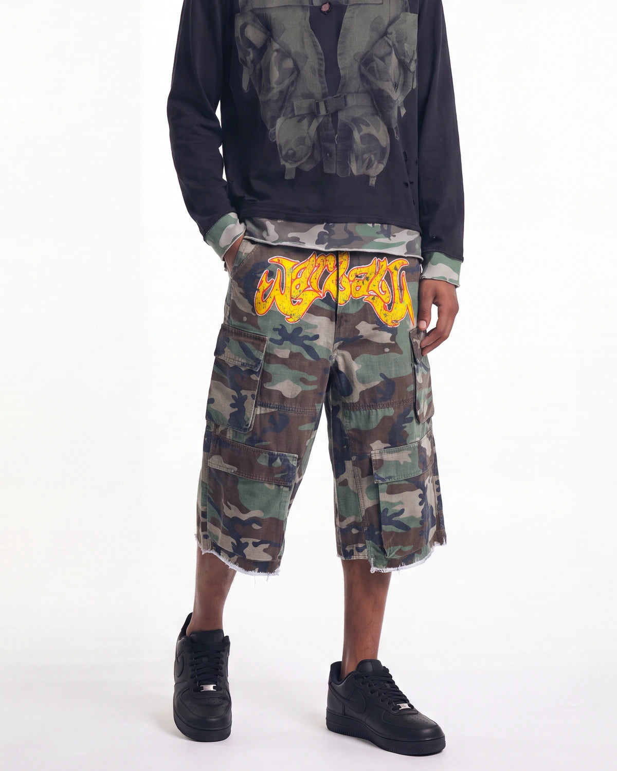A model wearing a pair of camouflage cargo shorts with a prominent yellow graphic print on the back waistband, styled with a black graphic long-sleeve shirt and black sneakers against a white studio background.