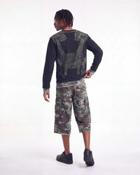 A model shown from the back wearing a black long-sleeve shirt with a green tactical vest graphic on the back, paired with green and brown camouflage cargo shorts and black shoes, against a plain white studio background.