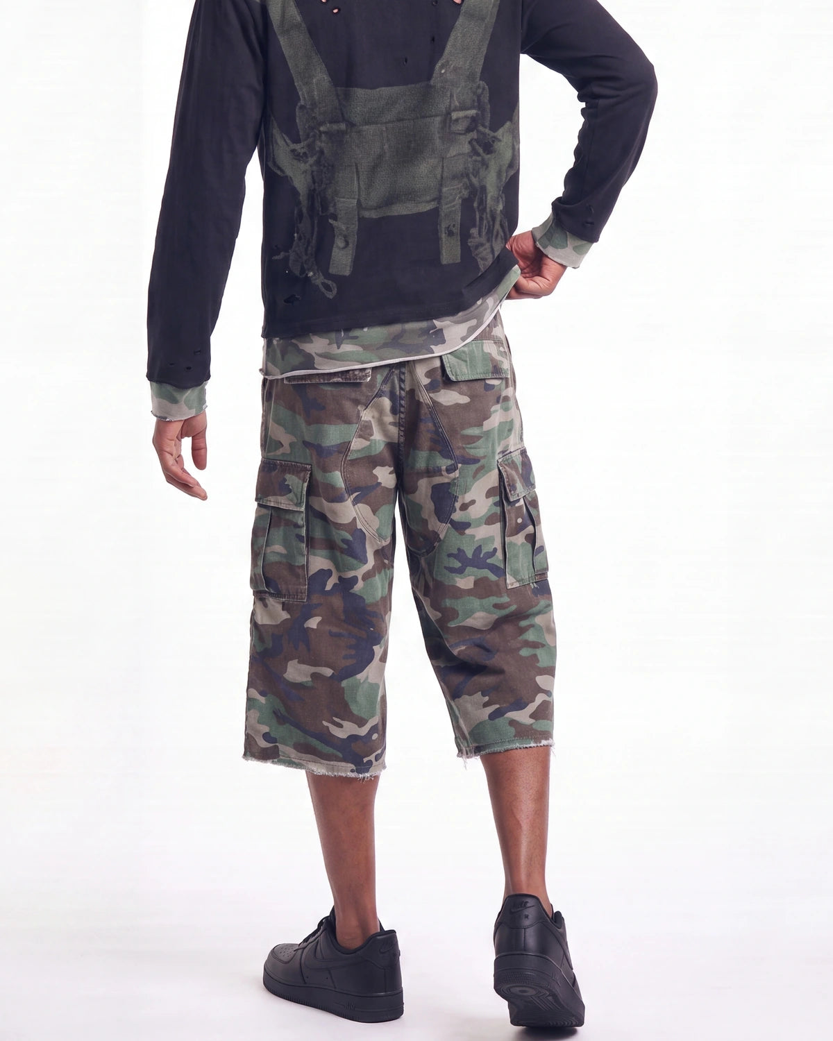 A model from the back wearing a black long-sleeve shirt with a green tactical harness graphic, green camouflage cargo shorts, and black shoes, against a plain white background.