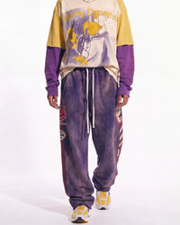 A model wearing a purple and yellow athletic outfit, featuring a long-sleeve graphic sweatshirt with a yellow and purple color-blocked design, matching purple sweatpants with white drawstrings and side patches, and yellow and white sneakers, against a plain white studio background.