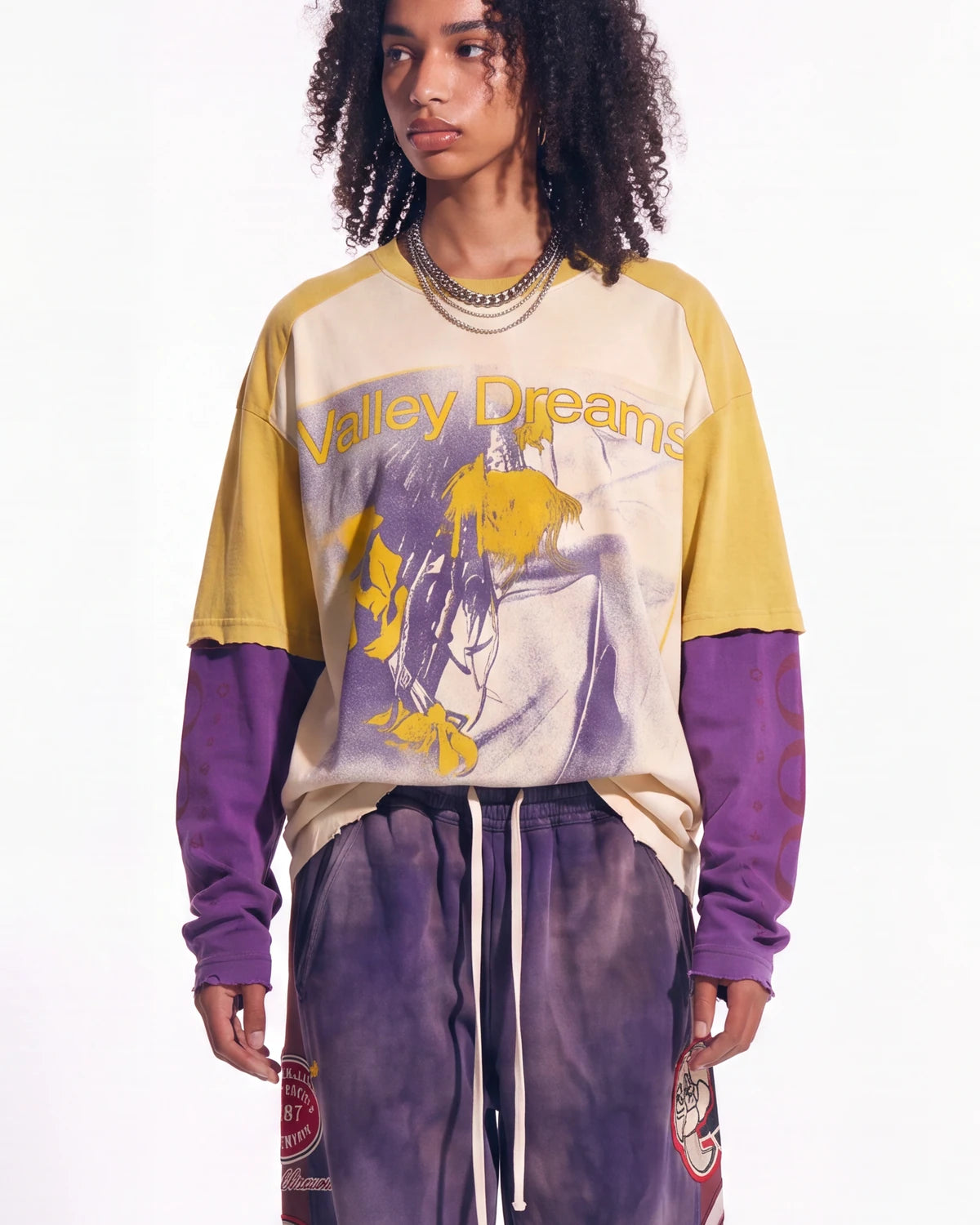 A model wearing a layered long-sleeve graphic t-shirt with a cream and yellow body, purple raglan sleeves, and a purple graphic print on the front reading 'Valley Dreams', paired with purple sweatpants against a white studio background.