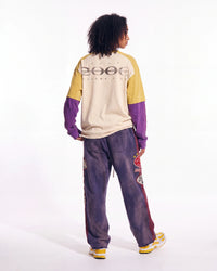 A model shown from the back wearing a color-blocked long-sleeve shirt with a '2006' graphic on the back, paired with matching purple tie-dye sweatpants and yellow sneakers, against a plain white studio background.