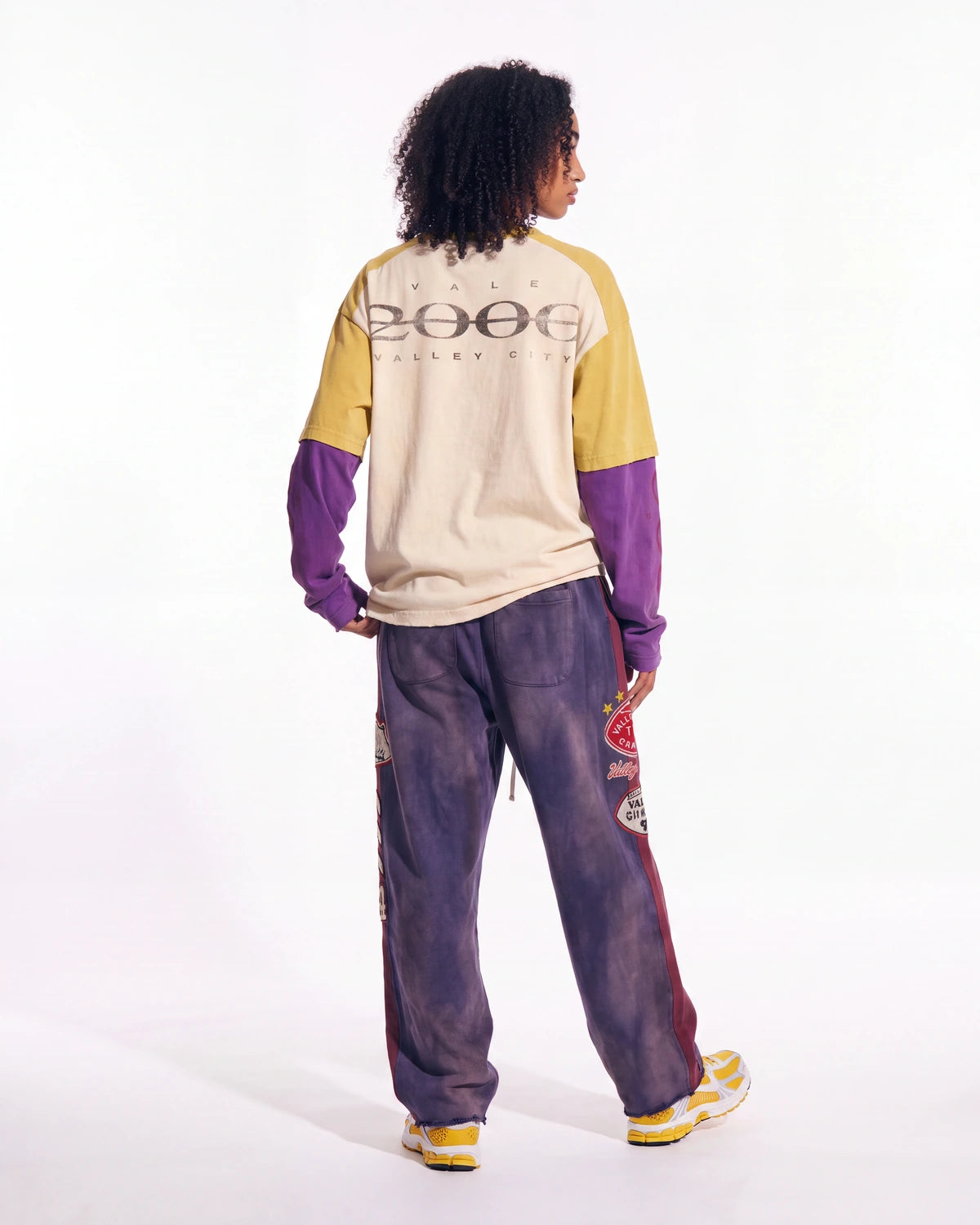A model shown from the back wearing a color-blocked long-sleeve shirt with a '2006' graphic on the back, paired with matching purple tie-dye sweatpants and yellow sneakers, against a plain white studio background.