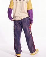 A person shown from the back wearing a colorblock long-sleeve shirt with a beige body, yellow shoulders, and purple sleeves, paired with purple sweatpants featuring red side panels and graphic patches, against a plain white studio background.