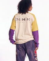 A model shown from the back wearing a color-blocked long-sleeve sweatshirt with a beige body, yellow shoulders, and purple sleeves, paired with purple sweatpants featuring graphic patches.