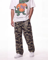 A person wearing camouflage cargo pants with a circular patch on the left thigh, styled with a white graphic t-shirt and white sneakers against a plain white background.