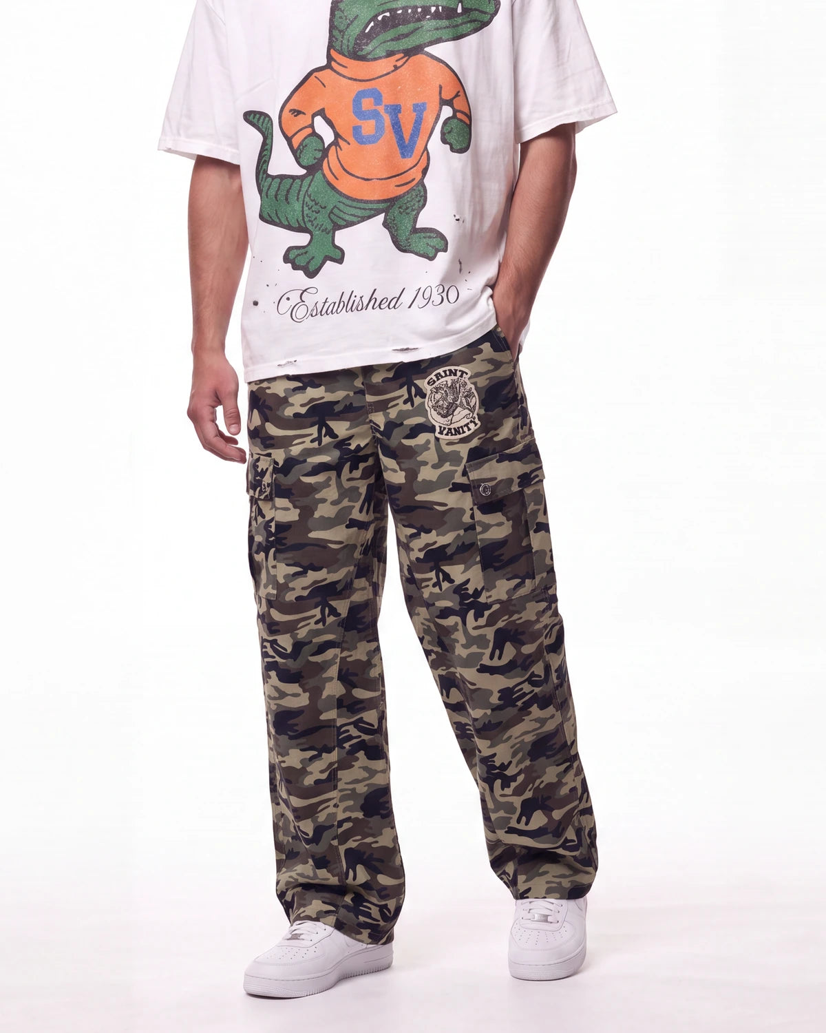 A person wearing camouflage cargo pants with a circular patch on the left thigh, styled with a white graphic t-shirt and white sneakers against a plain white background.