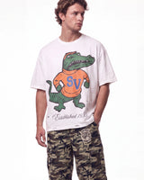 A man wearing a white graphic t-shirt with a green alligator mascot wearing an orange shirt with blue 'SV' lettering, paired with camouflage cargo pants, against a plain white background.