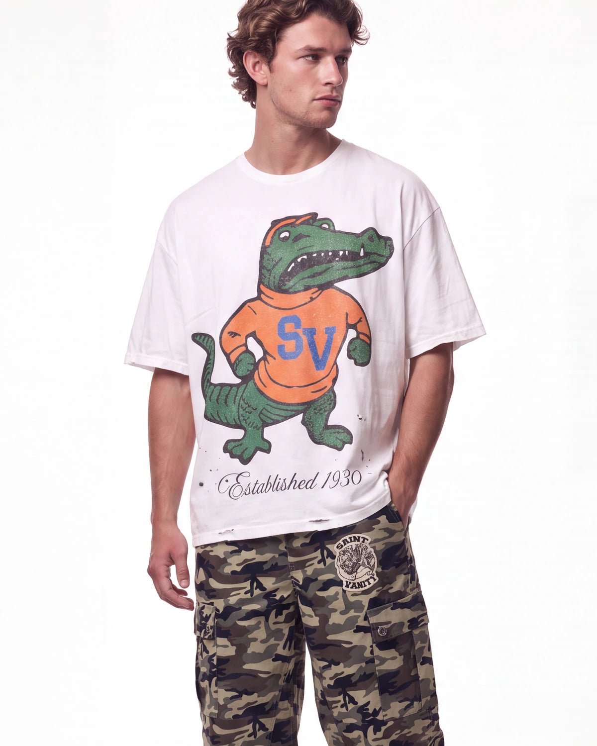 A man wearing a white graphic t-shirt with a green alligator mascot wearing an orange shirt with blue 'SV' lettering, paired with camouflage cargo pants, against a plain white background.