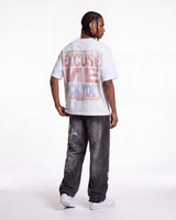 A man shown from the back wearing a white graphic t-shirt with a distressed red and blue text print that reads 'EXCUSE ME I'M NOT F*CK YOU', paired with dark gray washed jeans and white sneakers, against a plain white studio background.