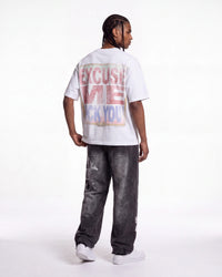 A man shown from the back wearing a white graphic t-shirt with a distressed red and blue text print that reads 'EXCUSE ME I'M NOT F*CK YOU', paired with dark gray washed jeans and white sneakers, against a plain white studio background.
