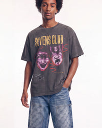 A man wearing a faded dark gray graphic t-shirt with 'SEVENS CLUB' and theater masks printed on the front, paired with blue jeans, against a plain white background.