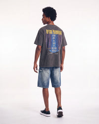 A man shown from the back wearing a faded dark gray graphic t-shirt with a blue and yellow print, paired with blue denim shorts and black sneakers, against a plain white studio background.