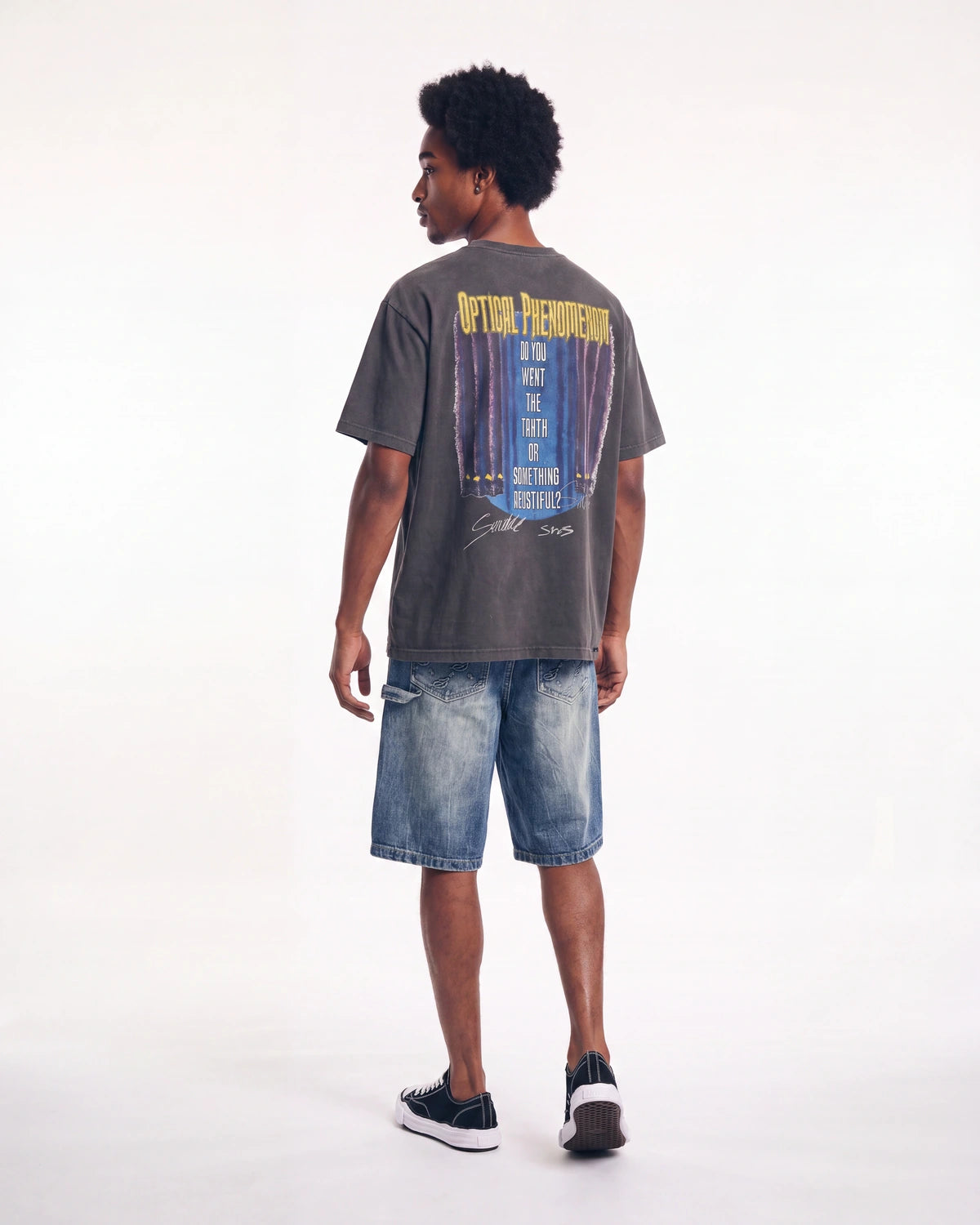 A man shown from the back wearing a faded dark gray graphic t-shirt with a blue and yellow print, paired with blue denim shorts and black sneakers, against a plain white studio background.