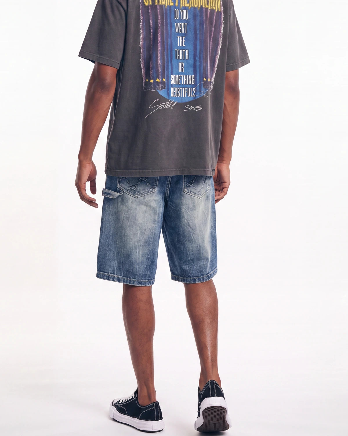 A model from the back wearing a faded black graphic t-shirt, blue denim shorts, and black low-top sneakers with white soles, set against a plain white studio background.