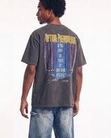 A man wearing a dark gray graphic t-shirt from the back, featuring a large print with yellow text and a blue curtain design. He is also wearing blue jeans, photographed against a plain white studio background.
