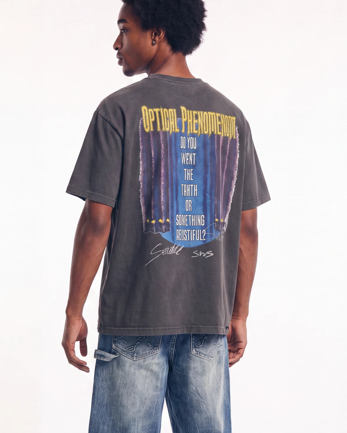 A man wearing a dark gray graphic t-shirt from the back, featuring a large print with yellow text and a blue curtain design. He is also wearing blue jeans, photographed against a plain white studio background.