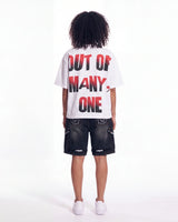 A child model shown from the back wearing a white t-shirt with the text 'OUT OF MANY, ONE' in red and black, paired with dark denim shorts and white sneakers, against a plain white studio background.
