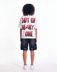 A child model shown from the back wearing a white t-shirt with the text 'OUT OF MANY, ONE' in red and black, paired with dark denim shorts and white sneakers, against a plain white studio background.