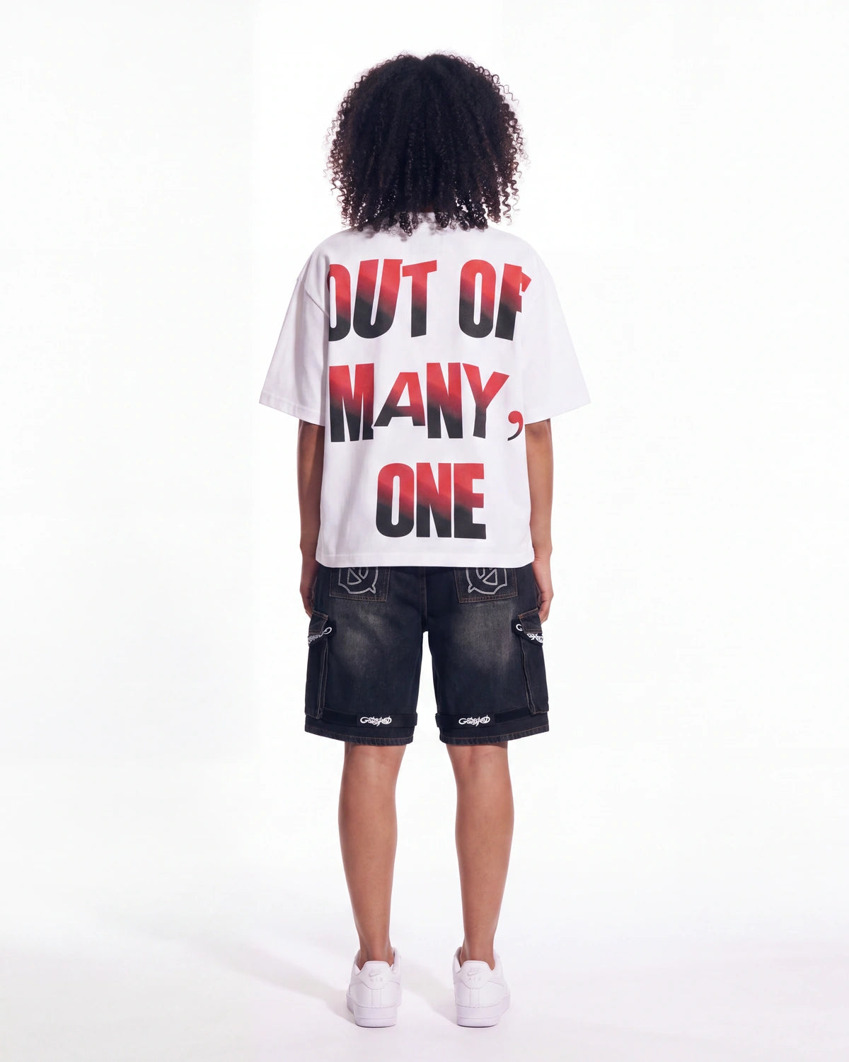 A child model shown from the back wearing a white t-shirt with the text 'OUT OF MANY, ONE' in red and black, paired with dark denim shorts and white sneakers, against a plain white studio background.