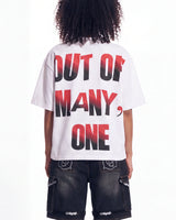 A person shown from the back wearing a white oversized T-shirt with large text reading 'OUT OF MANY, ONE' in black with a red gradient, paired with dark cargo shorts.