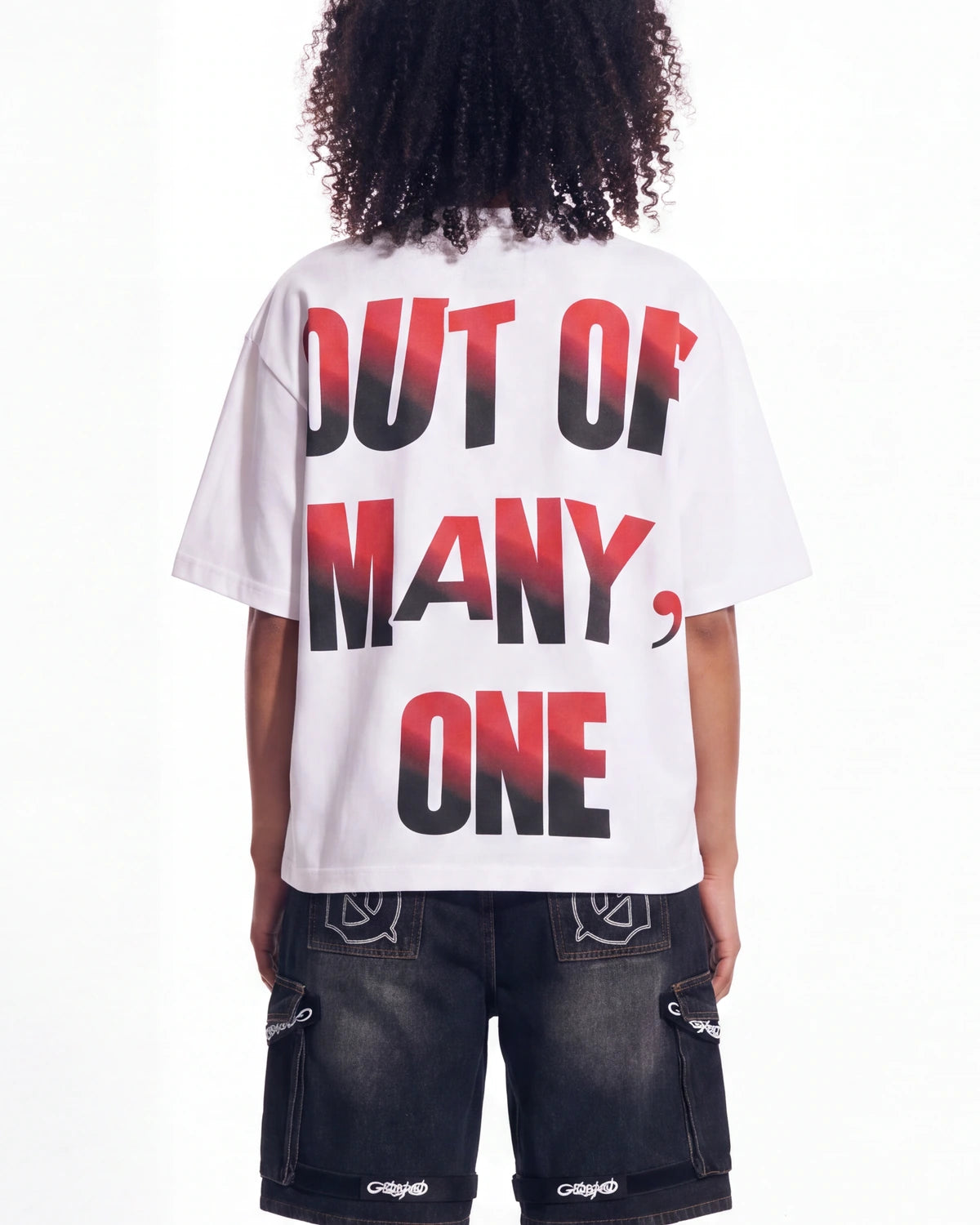 A person shown from the back wearing a white oversized T-shirt with large text reading 'OUT OF MANY, ONE' in black with a red gradient, paired with dark cargo shorts.