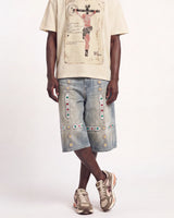 A model wearing light-wash denim shorts embellished with colorful rhinestones and gold-tone hardware, paired with a beige graphic t-shirt and New Balance sneakers, against a plain white studio background.