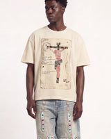 A man wearing a cream-colored t-shirt with a large vintage-style crucifix graphic print on the front, paired with light-wash denim jeans featuring colorful embroidered details, set against a plain white studio background.