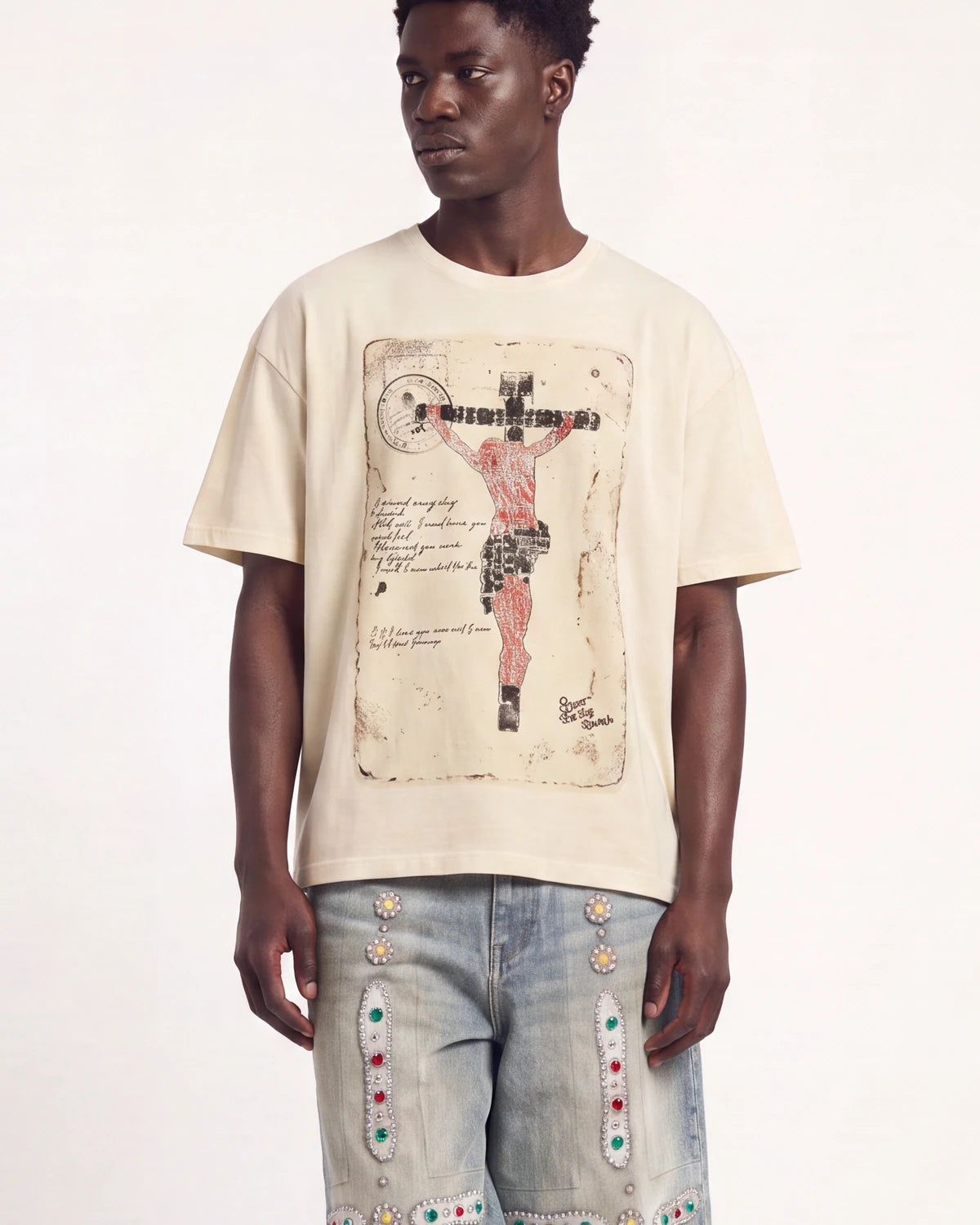 A man wearing a cream-colored t-shirt with a large vintage-style crucifix graphic print on the front, paired with light-wash denim jeans featuring colorful embroidered details, set against a plain white studio background.