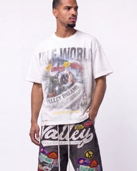 A man wearing a white graphic t-shirt with a distressed 'Valley World' print and black pants covered in colorful patches, standing against a plain white studio background.