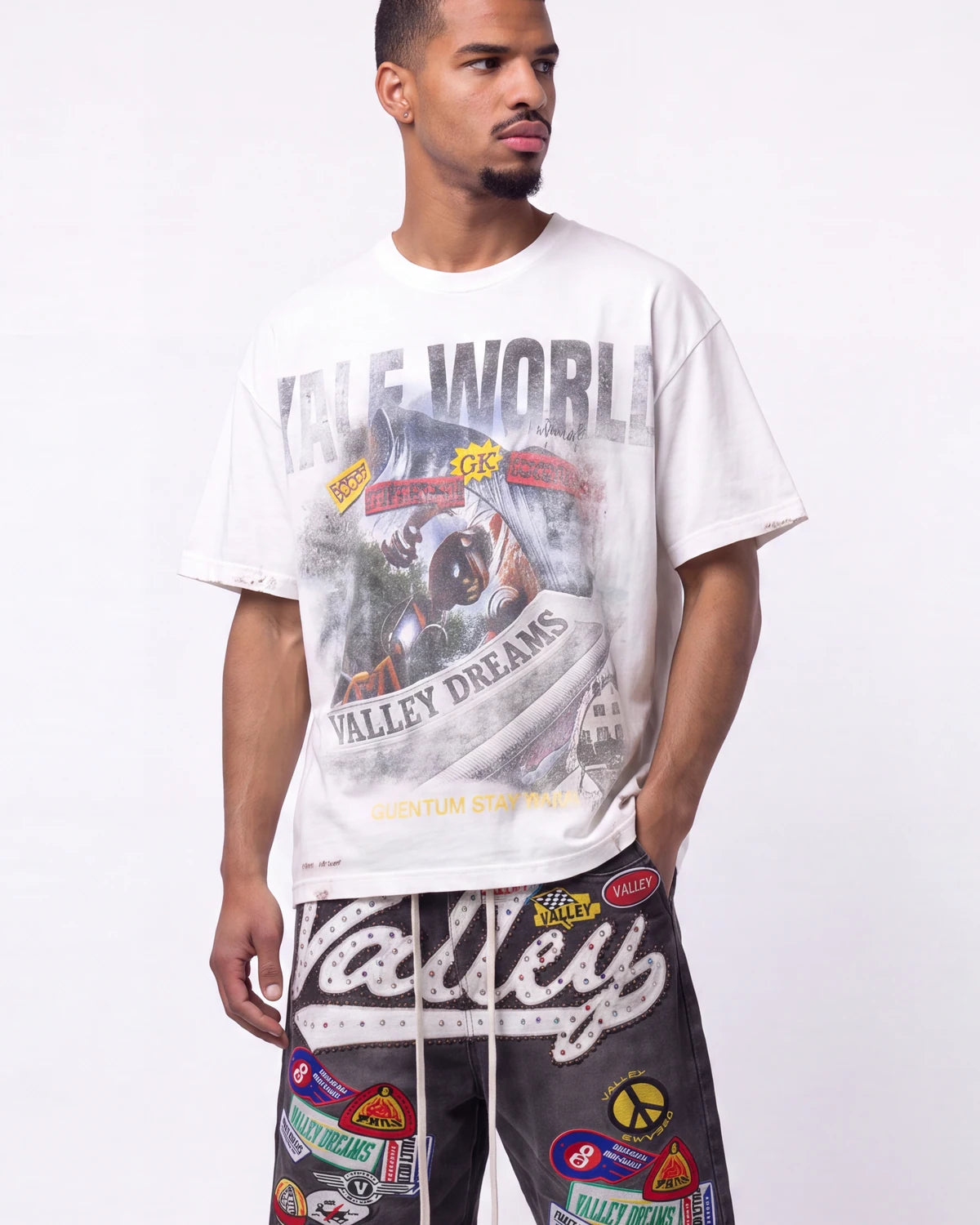 A man wearing a white graphic t-shirt with a distressed 'Valley World' print and black pants covered in colorful patches, standing against a plain white studio background.