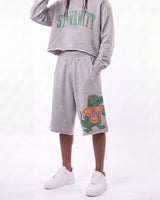 A person wearing a light gray distressed sweatshirt with green 'ST. VANITY' lettering and matching light gray sweat shorts featuring a green alligator graphic with an orange 'SV' shirt, paired with white sneakers against a white studio background.
