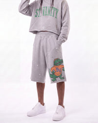 A person wearing a light gray distressed sweatshirt with green 'ST. VANITY' lettering and matching light gray sweat shorts featuring a green alligator graphic with an orange 'SV' shirt, paired with white sneakers against a white studio background.