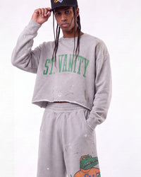 A model wearing a light gray distressed crewneck sweatshirt with 'ST. VANITY' printed in green across the chest, matching sweatpants with a green alligator graphic near the hem, and a black baseball cap.